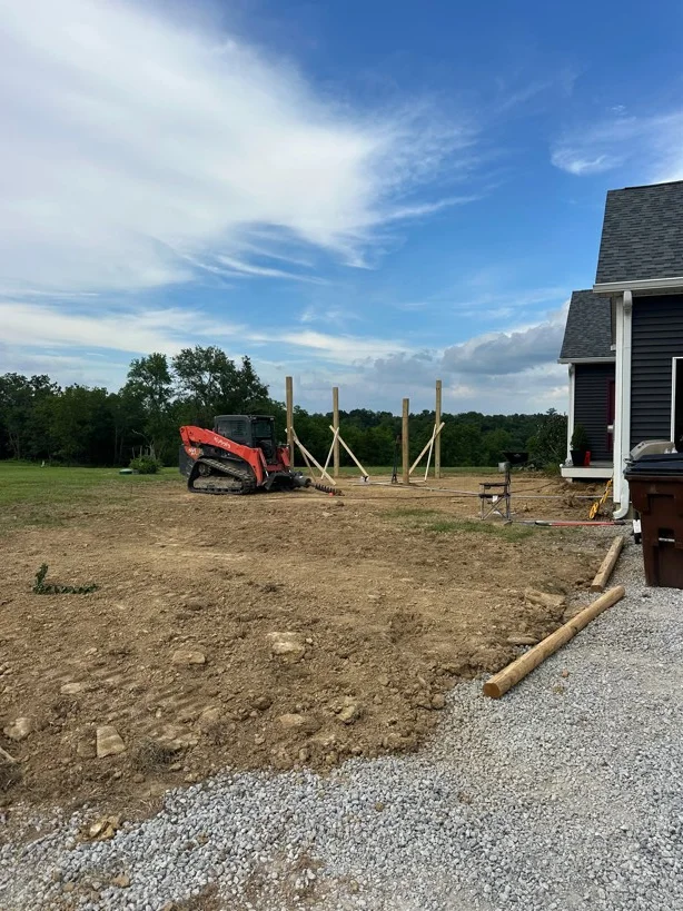 Excavated residential lot with graded soil, wooden post footings, and gravel driveway approach