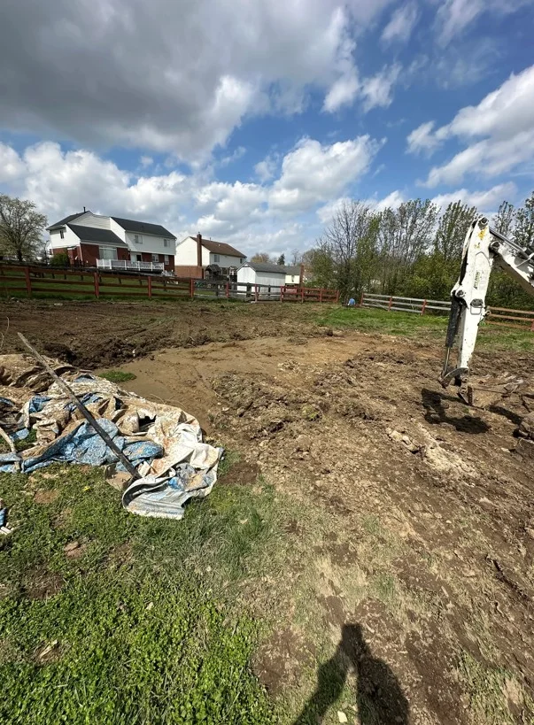 Excavated residential lot with mini excavator, cleared soil, and wooden fence perimeter